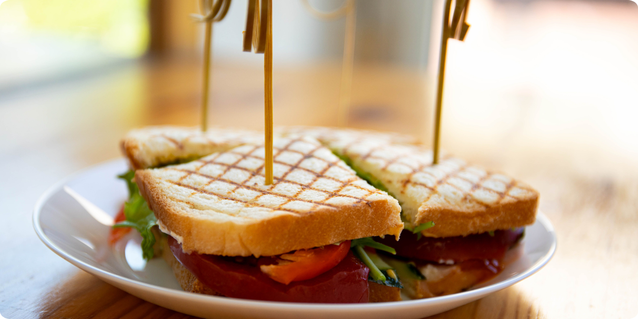 A freshly made sandwich on a white plate, garnished with lettuce and tomato, placed on a wooden table