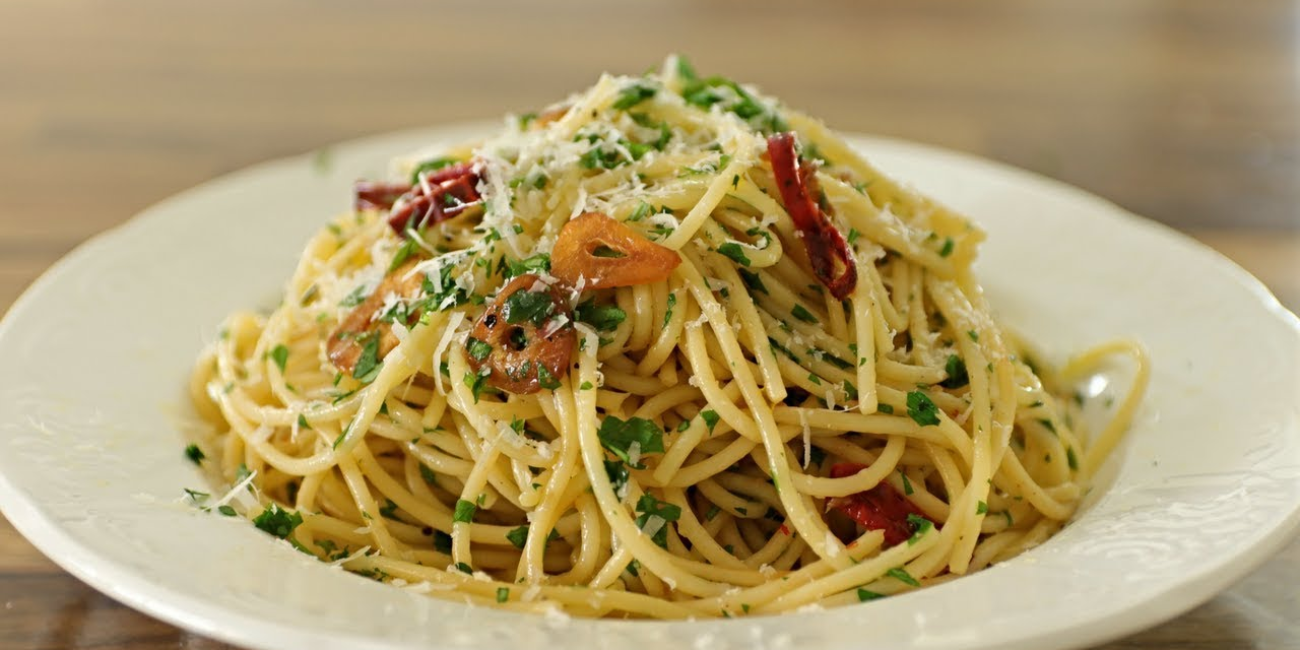 A plate of Spaghetti Aglio e Olio topped with fresh tomatoes and herbs, served with a side of chicken and vegetables