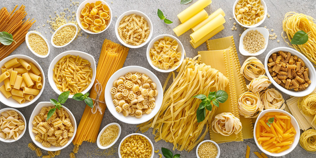 Assorted uncooked pasta varieties are arranged in bowls and on a gray surface, with some basil leaves scattered among them