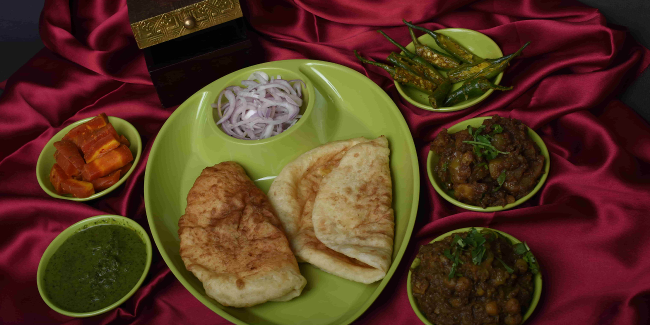 A green plate with two pieces of bhature and sliced onions, surrounded by small bowls containing green chutney, chopped carrots, green chilies, and two different chole on a red satin fabric.