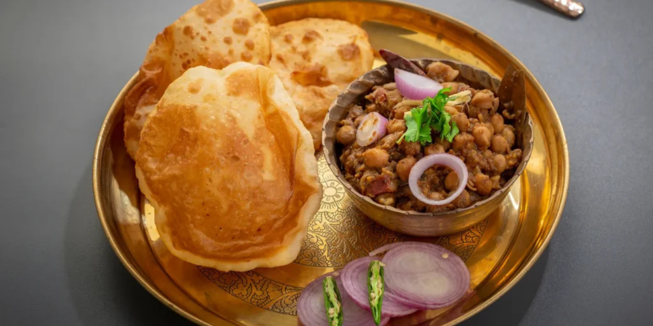 Golden Indian thali with bhature and a bowl of spicy chole topped with onion and cilantro, served with sliced onion and green chilies. аппetizing meal.